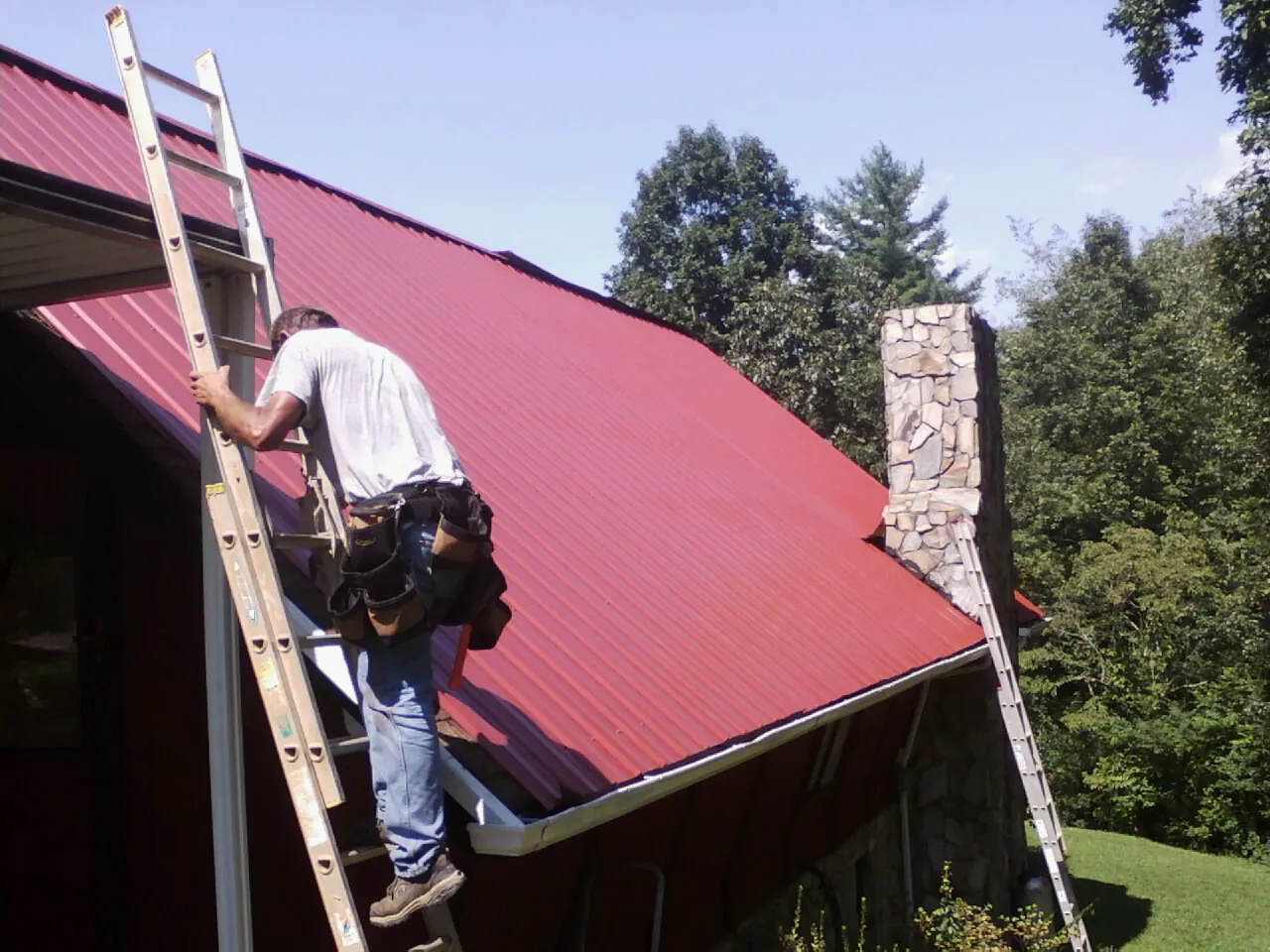 Roofer on a ladder beside a red metal roof and stone chimney — Soffit & Fascia Repair by Roofing Brothers in Kenneth